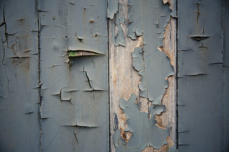 Rotting wood siding with gray paint peeling from the panels.