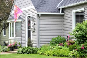 A gray home with black asphalt shingle roofing and a green garden around the home.