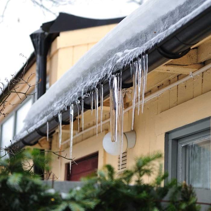 Snow and icicles hanging from the roof of a yellow home with black gutters.