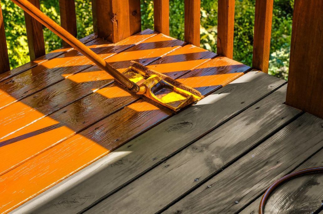 Staining wooden deck with paint roller; untreated patch of wood shown for contrast.