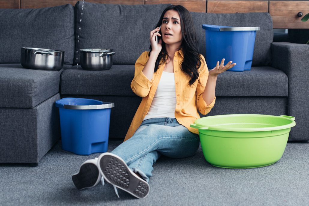 Worried woman talking on smartphone and sitting on floor under leaking ceiling in living room
