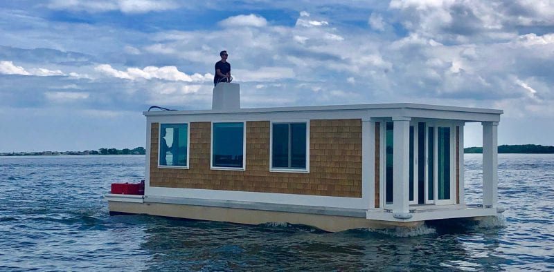 A house boat on a lake with tan Beach House shake siding and white trim and windows.