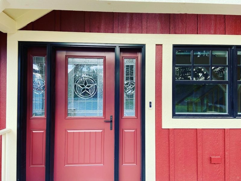 A red fiberglass entry door with decorative glass and black trim on a home with red board & batten siding.