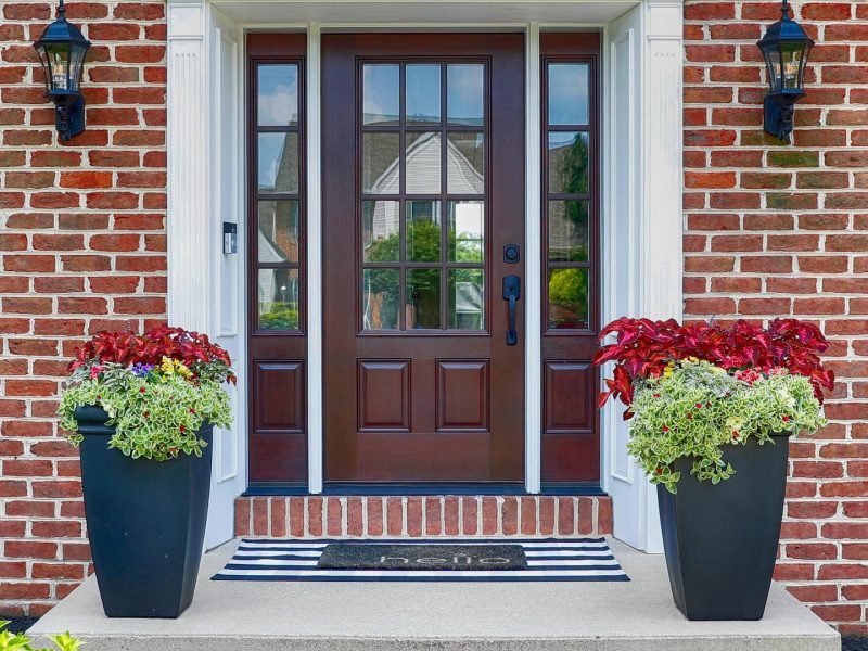A mahogany wood entry door on a brick house with floral planters on either side of the door.