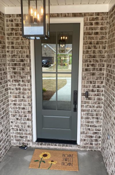 A gray entry door on a brick home with a door mat that reads home sweet home.